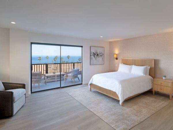 Hotel room with a large bed, nightstands, a sitting area, and a balcony overlooking the ocean through a sliding glass door. End.