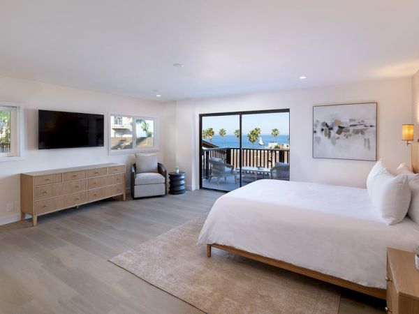 A bright hotel room with a king bed, a dresser and TV, a seating area, and a balcony overlooking water, airy and modern.