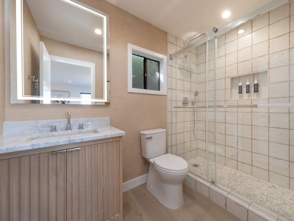 Modern bathroom with a double vanity, large backlit mirror, beige walls, and a glass-enclosed shower with tiled walls and built-in niches.