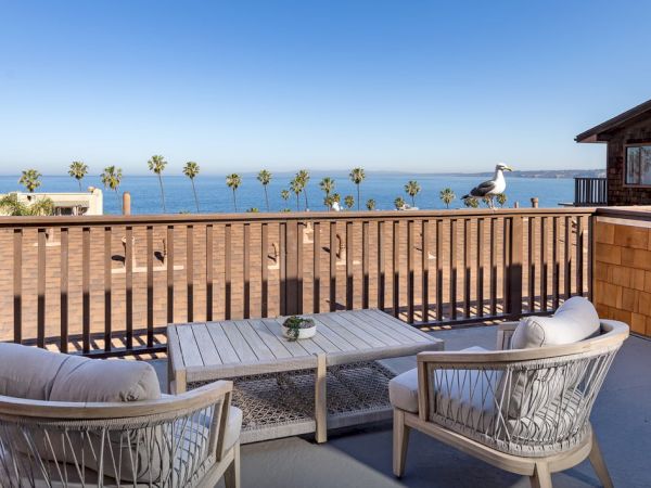 A seaside deck with wicker furniture overlooking a calm ocean, palm trees lining the horizon, and a cozy sitting area under a clear blue sky.