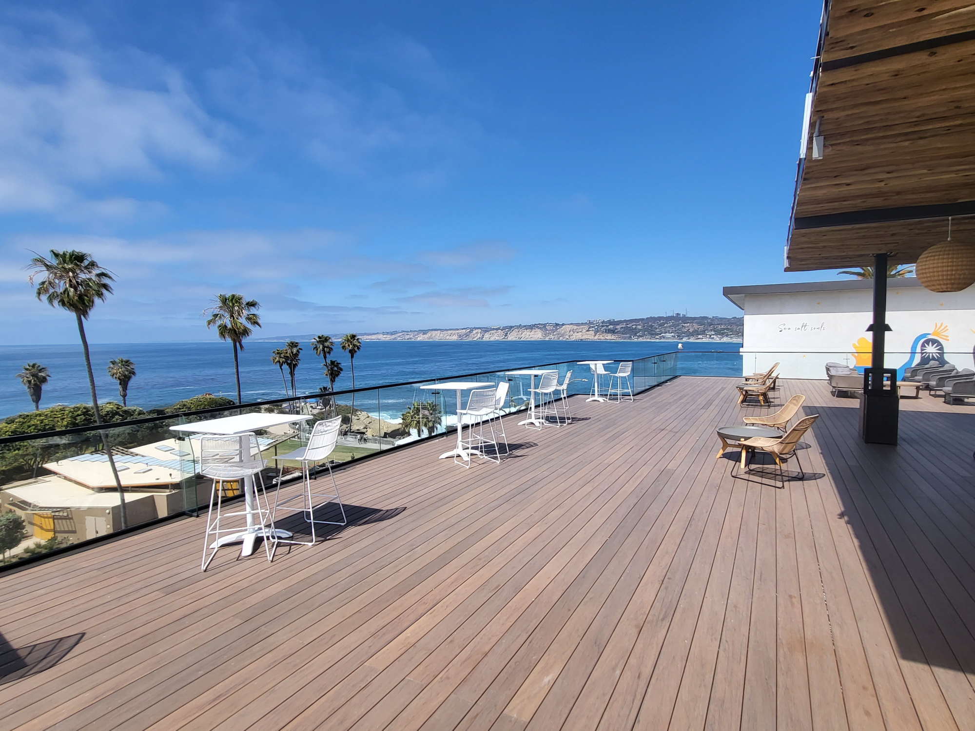 A sunny oceanfront deck with wooden flooring, white tables and chairs, palm trees, blue sea, and a clear sky, overlooking the coastline.