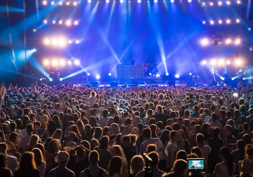 A large concert crowd facing a brightly lit stage with performers and speakers; colorful stage lights and screens illuminate the audience.