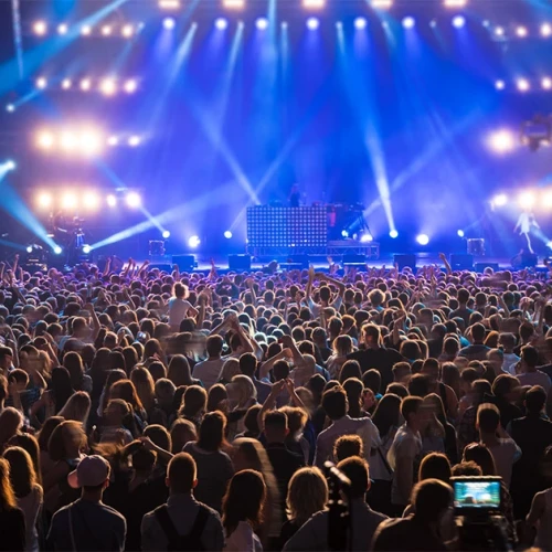 A large concert crowd facing a brightly lit stage with performers and speakers; colorful stage lights and screens illuminate the audience.
