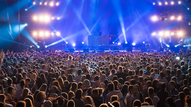 A large concert crowd facing a brightly lit stage with performers and speakers; colorful stage lights and screens illuminate the audience.