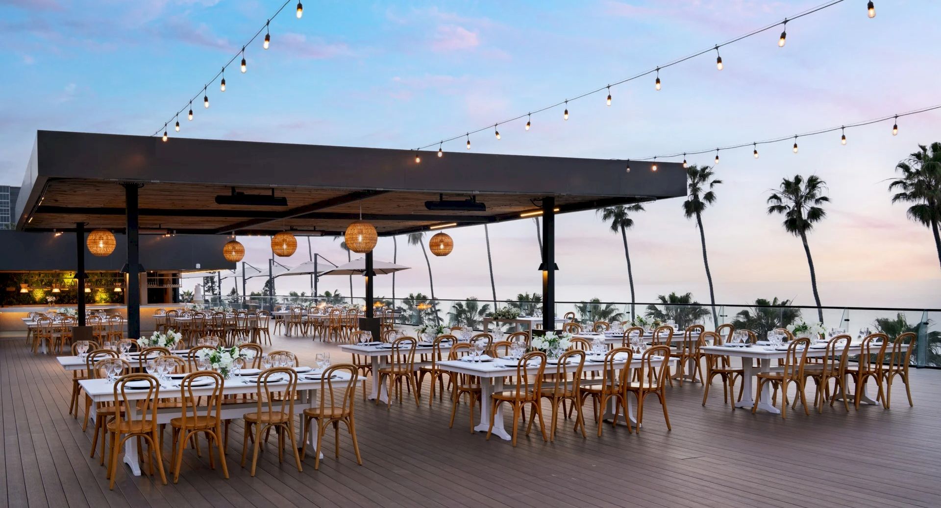 An outdoor beachfront dining setup with many wooden tables and chairs under a covered area, string lights, palm trees, and a sunset sky.